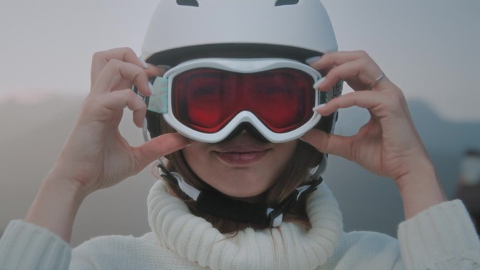 一个漂亮的白人女孩把一个白色的滑雪头盔或滑雪板头盔戴在头上。一个漂亮女人在冬季度假胜地的肖像。戴上防
