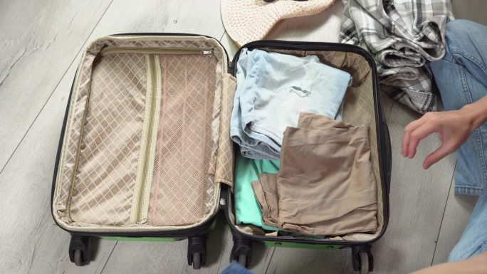 一个人把他的手提箱装上一叠钱准备长途旅行。盖上绿色的行李箱，手提行李里的衣服拉上拉链。旅游者为旅行打