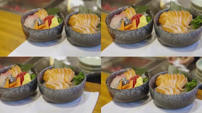 在一家生鱼片餐厅，厨师将三文鱼和什锦鱼生鱼片碗放在服务柜台上准备上桌，并在厨房柜台后面继续烹饪。