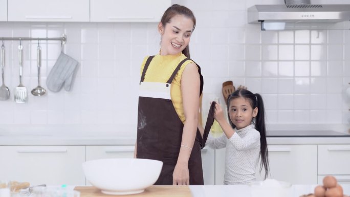 亚洲小女孩在现代厨房里帮妈妈系围裙，同时做面包房的照片。父母在假期一起做饭。