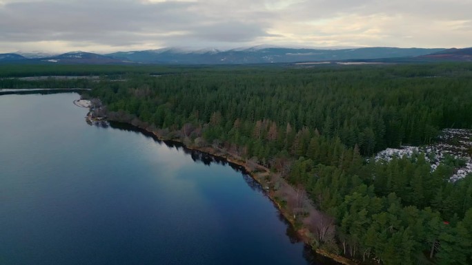 湖周围的树木的空中圆形镜头。平静的水面反射着岸边的树木。远处的山被雪覆盖。英国苏格兰