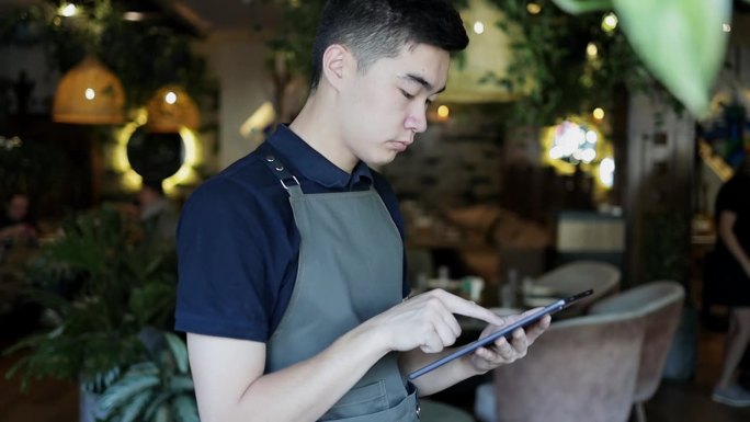严肃的咖啡厅员工穿着墨绿色的围裙，通过便携式平板电脑在网上点餐。穿着制服的有魅力的餐厅经理通过互联网