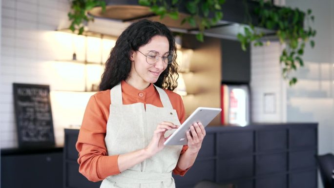 一名年轻的女小企业主站在当地一家咖啡馆里，手里拿着平板电脑，看着镜头
