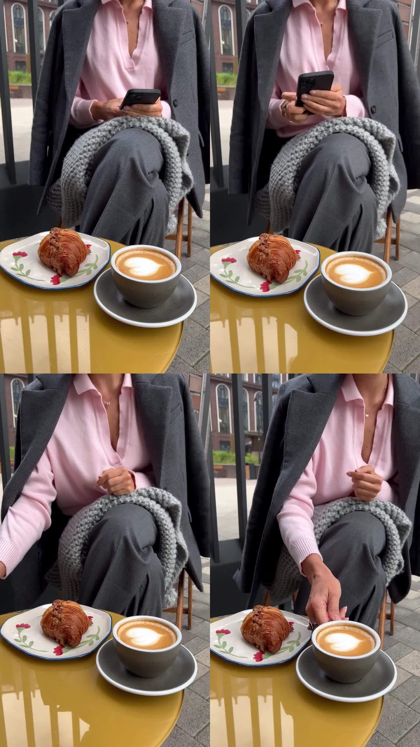 坐在户外咖啡馆里的女人，一边浏览智能手机，一边发信息，手里端着一杯卡布奇诺咖啡，桌上放着灰色格子围巾
