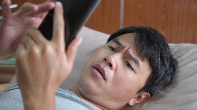 亚洲成年男子拿着智能手机或手机看得很紧
在卧室的床上皱着眉头。技术、通信、数字连接的概念。
