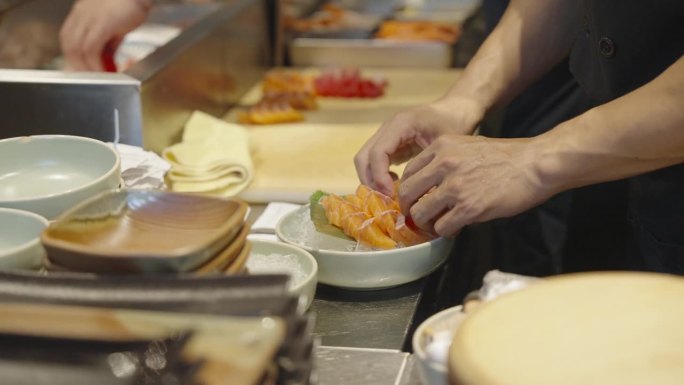 在一家日本餐馆里，一位厨师正在用手把三文鱼片放在冰盘里，另一位厨师正在给鱼切鱼片。