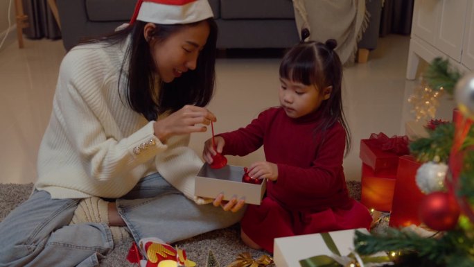 亚洲年轻快乐的家庭晚上在家里一起享受新年假期聚会。妈妈和孩子一起把礼品盒放在圣诞树旁装饰。度假的生活