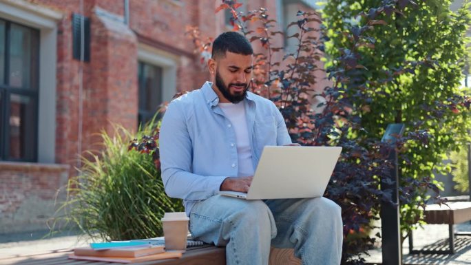 英俊的阿拉伯人坐在木凳上，拿着咖啡和书，在现代无线笔记本电脑上工作。穿着随意的年轻男性感觉疲惫，偏航