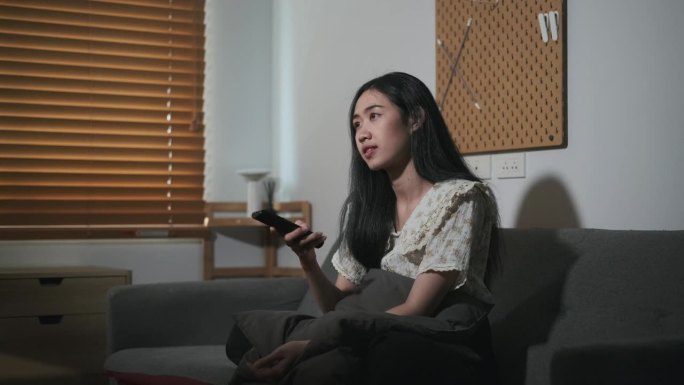 年轻的亚洲女人拿着遥控器切换频道，晚上在家看电影。