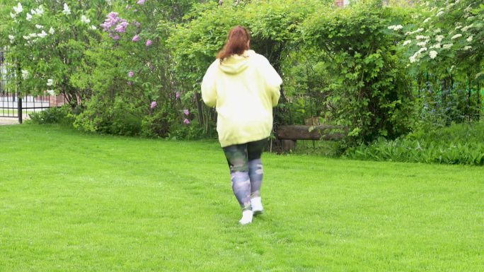 后视镜:身材臃肿的卷发女人穿着黄色连帽衫，做有氧运动，在公园里跑步