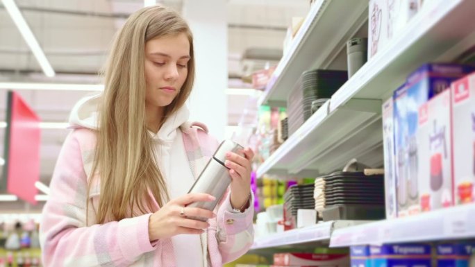 感兴趣的年轻女子在超市橱窗旁挑选保温瓶。