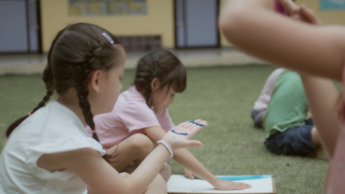 在一所户外小学，一位亚洲老师正在给可爱的男孩和女孩们在纸上画水彩画。