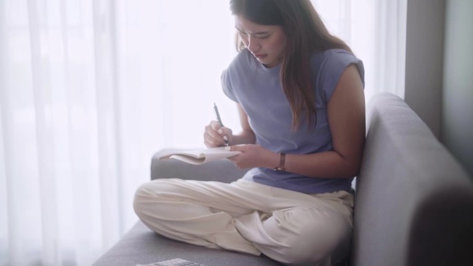 年轻的亚洲女性坐在家里客厅的沙发上，用计算器计算收入和支出或管理每月的预算，并把它写在笔记本上。