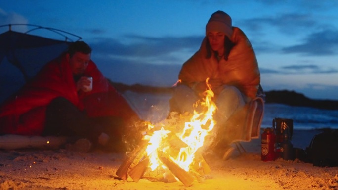 一对浪漫的夫妇穿着暖和的衣服在寒冷的天气里露营。一起在沙滩上篝火边热身，喝着热茶