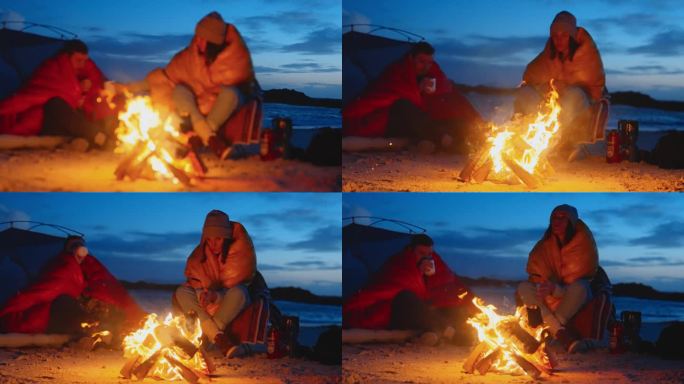 一对浪漫的夫妇穿着暖和的衣服在寒冷的天气里露营。一起在沙滩上篝火边热身，喝着热茶