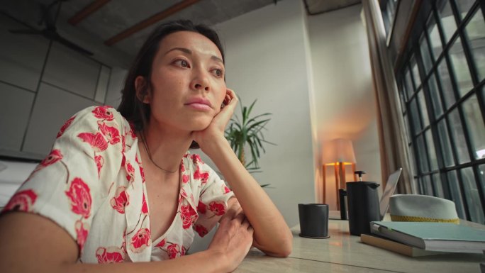 亚洲女性对创造性工作的思考或对家庭办公室室内规划的考虑。忧郁的女士看起来很紧张，坐在房间的桌子旁。世