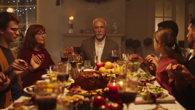 一位老人与家人和孩子坐在节日餐桌的首位，在吃火鸡晚餐前祈祷。宗教男性祝福圣诞食品，感谢上帝和耶稣基督