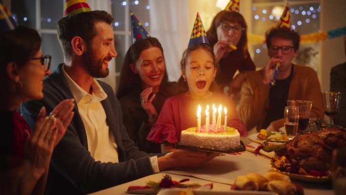 年轻英俊的父亲给他可爱的女儿带了一个带蜡烛的生日蛋糕。兴奋的女孩在家里的家庭晚宴上吹蜡烛并许愿
