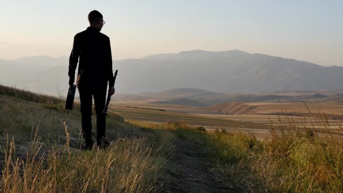一个年轻的音乐家，穿着黑色的衣服，手拿黑色的单簧管和一个乐器的袋子，在夕阳的山间，沿着道路走向远方。