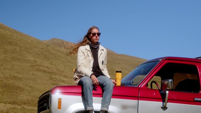迷人的长发女孩站在一辆老式汽车旁边，以山景为背景，用保温杯喝着饮料。