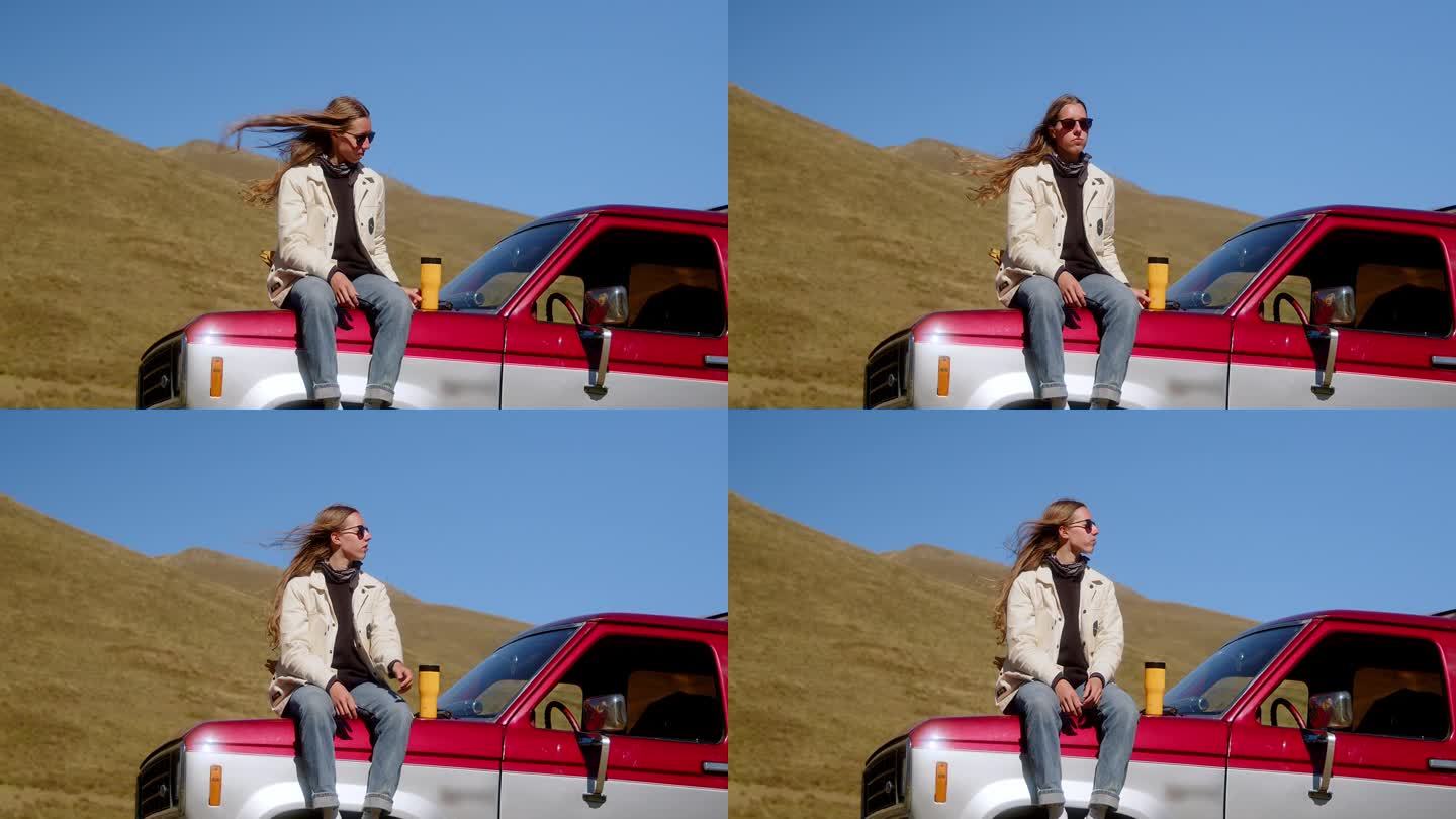 迷人的长发女孩站在一辆老式汽车旁边，以山景为背景，用保温杯喝着饮料。