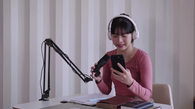 快乐的亚洲妇女设置播客工作室戴着耳机，用麦克风录制播客，在智能手机上阅读新闻，在家庭工作室进行广播在