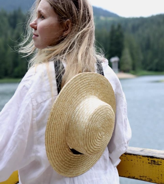 一个穿着白衬衫，肩上戴着草帽的女孩站在湖边的群山中。一位美丽的游客正在山里旅行。垂直视频