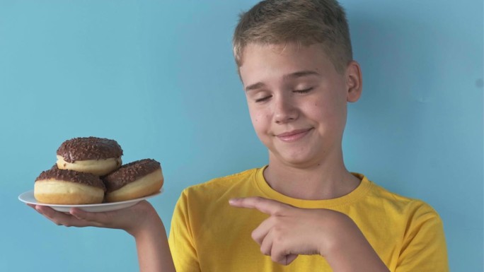 快乐可爱的少年指着蓝色背景的满满一盘巧克力甜甜圈。传递和不健康甜食的概念。