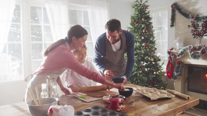 圣诞快乐的家庭:年轻的父母和他们可爱的小女儿在家里一起准备糕点的慢动作肖像。可爱的家庭准备节日晚餐，