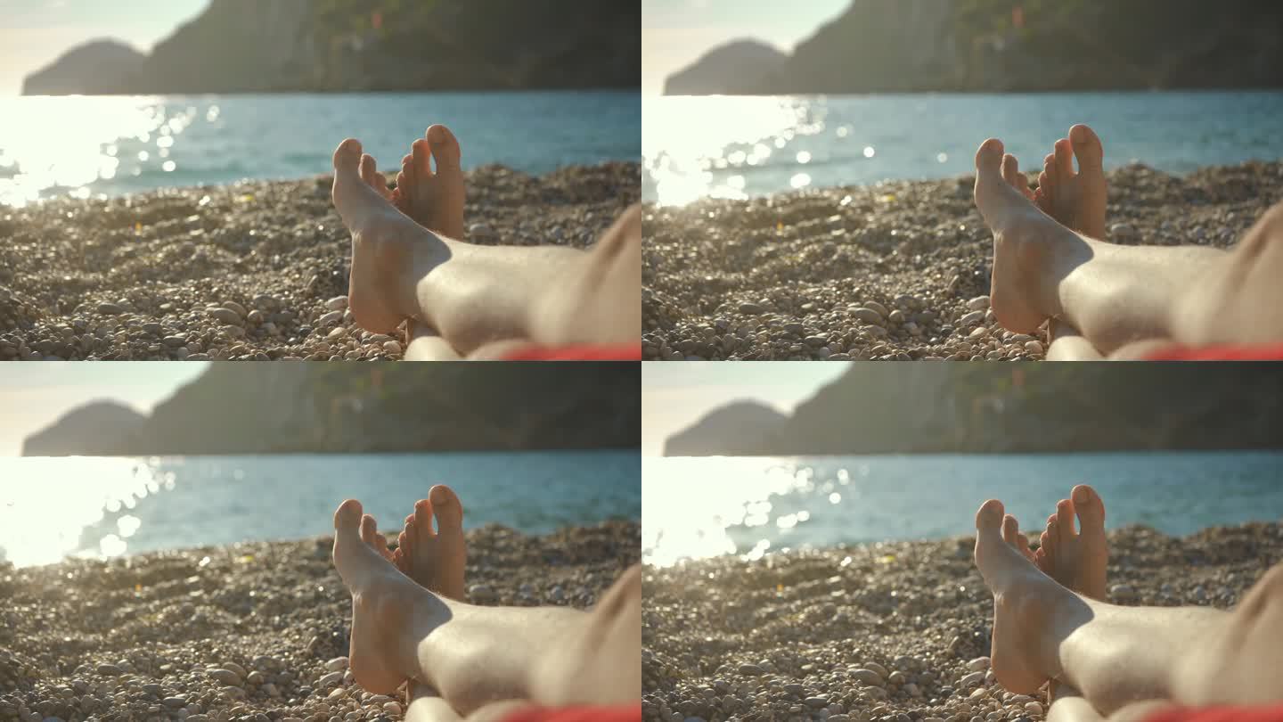 年轻人躺在卵石滩上休息，只看得见交叉的光脚，迷离的午后阳光照亮了平静的海面，背景是岩石嶙峋的悬崖