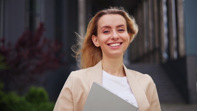 自信迷人的女商人的肖像，女人看着镜头微笑着拿着笔记本电脑，风吹着头发，骄傲的企业家在外面享受成功的创