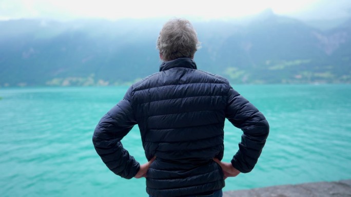 背影成熟的男子站在细雨中凝视着湖光山色的美景，悠闲的人穿着夹克在平静的沉思中