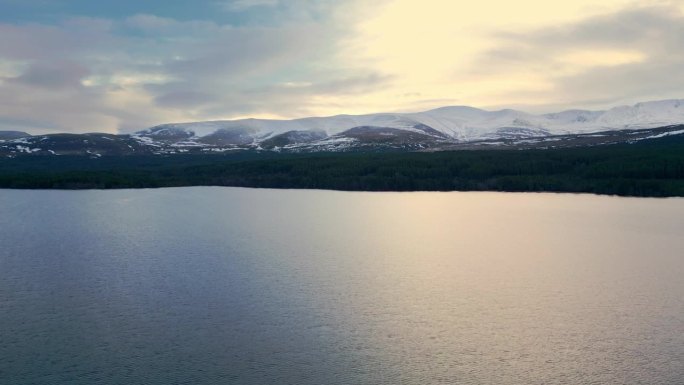 在冬季的高山景观中，向前飞越湖面。黄昏时多云天空下白雪皑皑的山脊。英国苏格兰