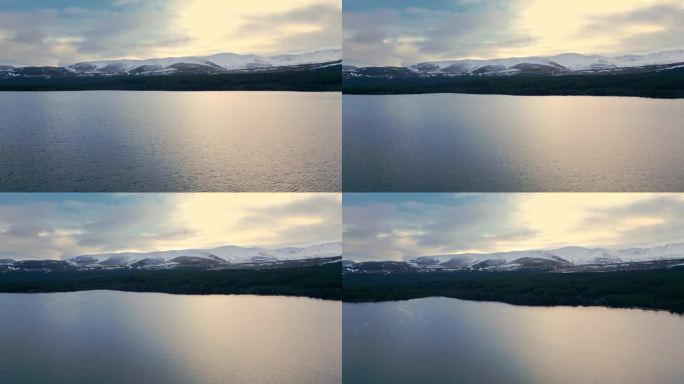 在冬季的高山景观中，向前飞越湖面。黄昏时多云天空下白雪皑皑的山脊。英国苏格兰