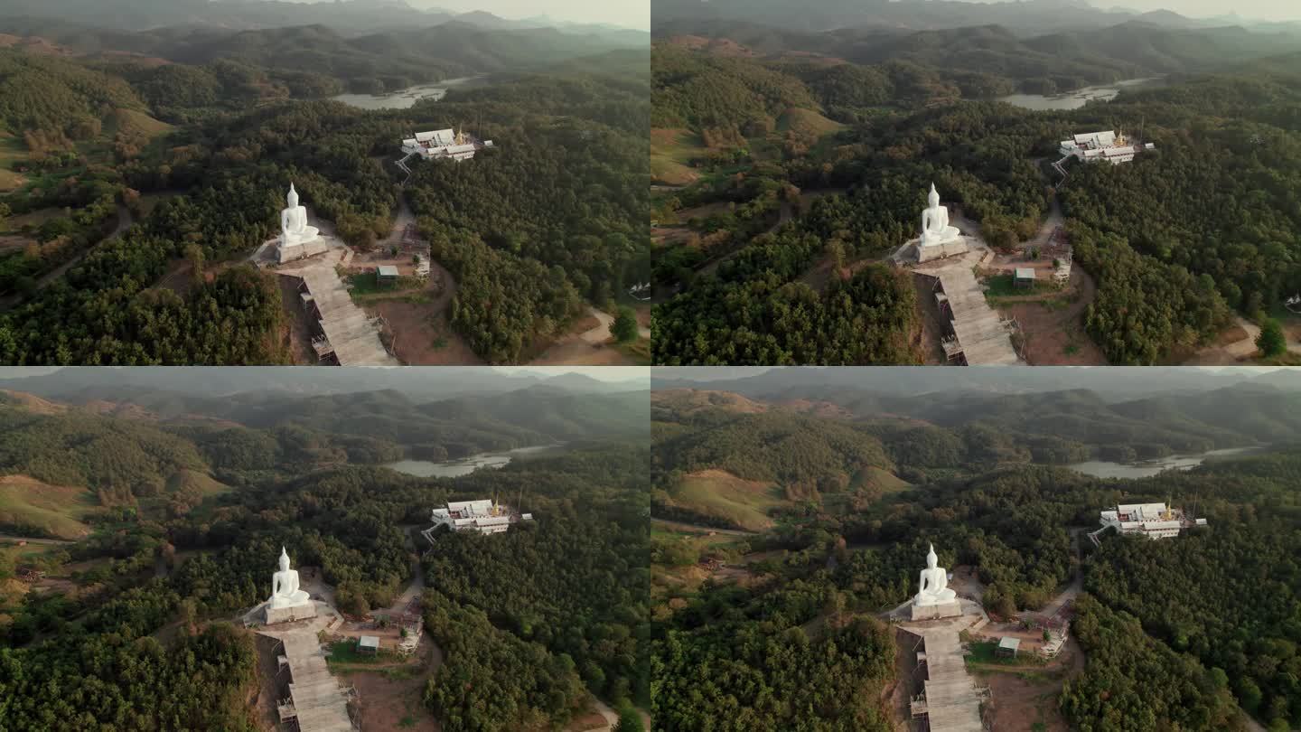 在一个阳光明媚的日子里，乡村树林环绕的山顶上，有金色宝塔、白色小教堂和白色佛像的佛教寺庙的鸟瞰图