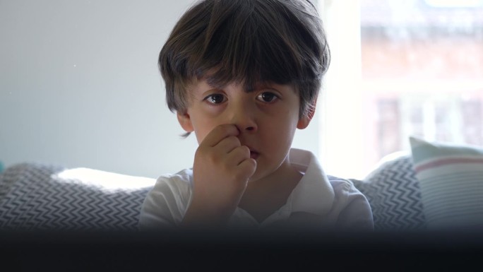 小男孩盯着电视屏幕挖鼻孔的特写，孩子沉浸在家里的娱乐中