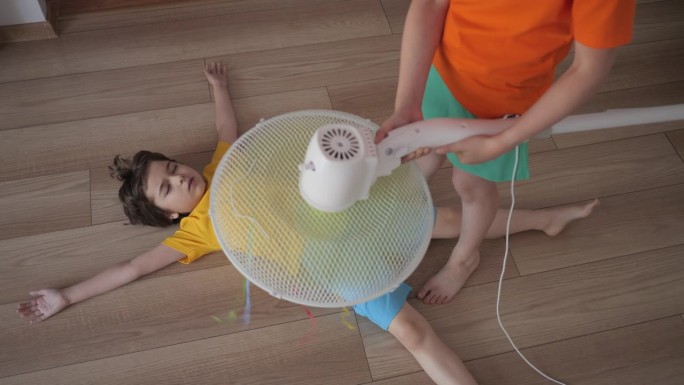 男孩和女孩在扇子前大热天在家里客厅。炎炎夏日，孩子们在家中享受电风扇吹来的凉风。酷暑的孩子们享受着吹