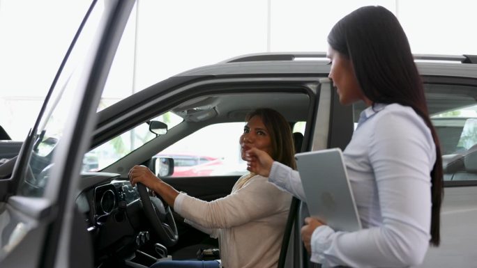 漂亮的女售货员向坐在驾驶座上的成熟女顾客展示汽车的内饰，看起来很开心