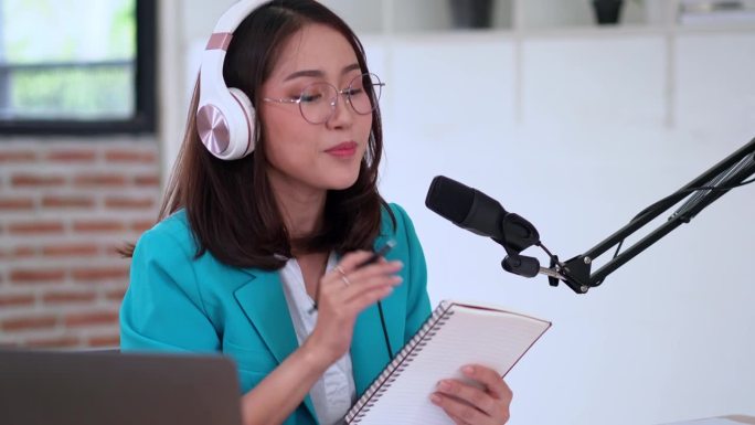 年轻漂亮的亚洲女性dj对着麦克风讲话，录制播客，从记事本上读笔记，或者在录音棚里做新闻广播