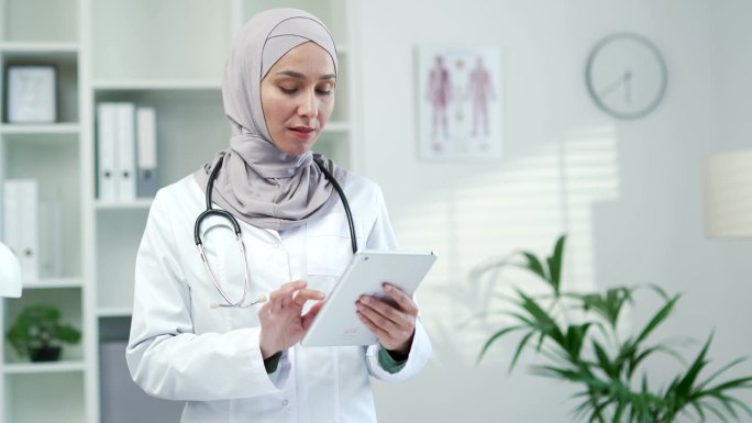 在一家现代医院的诊所里，身穿白大褂的穆斯林女医生正在平板电脑上使用浏览式打字
