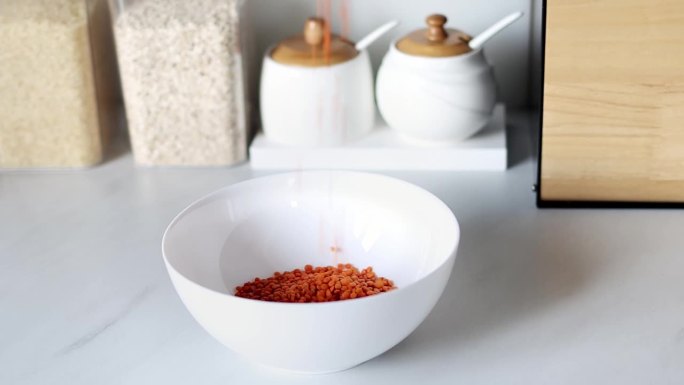 红色的扁豆被倒入白色的盘子里，背景是现代厨房的慢动作。健康食品和营养理念。安全、生态、健康产品