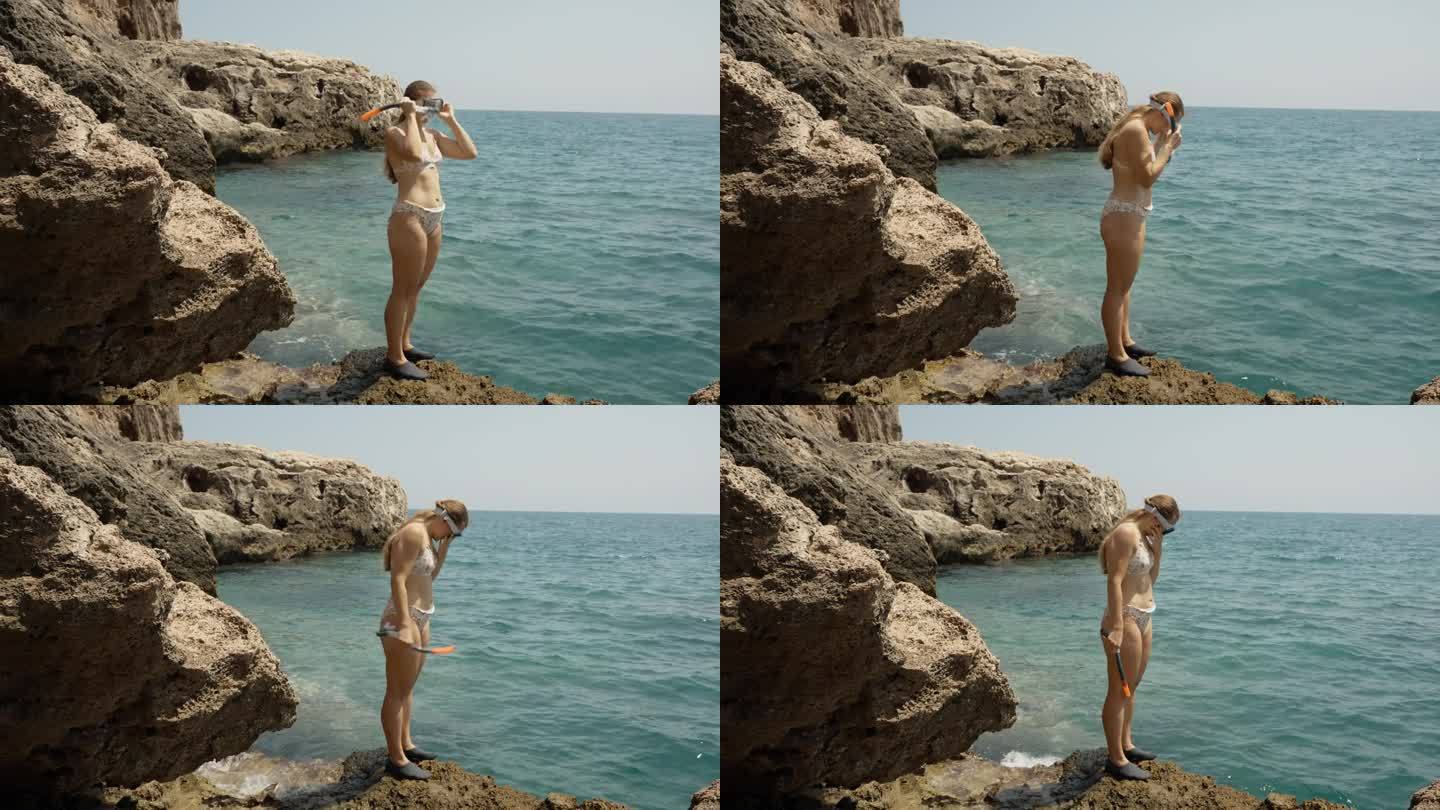 一个穿着泳装的年轻女子站在海边的悬崖边上，戴上带呼吸管的潜水面罩。