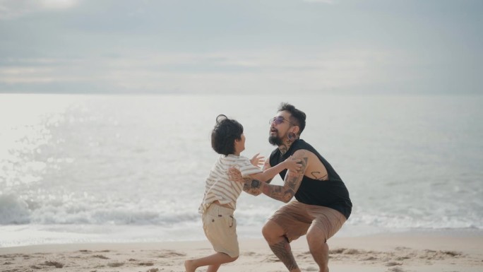 一位亚洲父亲在沙滩上以美丽的天空为背景，抱着年幼的儿子围成一圈。
