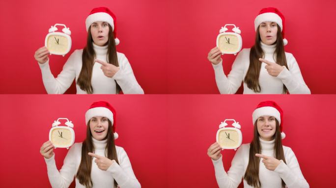 快乐的年轻女子穿着暖和的毛衣，戴着圣诞老人帽，用食指指着5点到12点的时钟，在淡红色的工作室背景下独