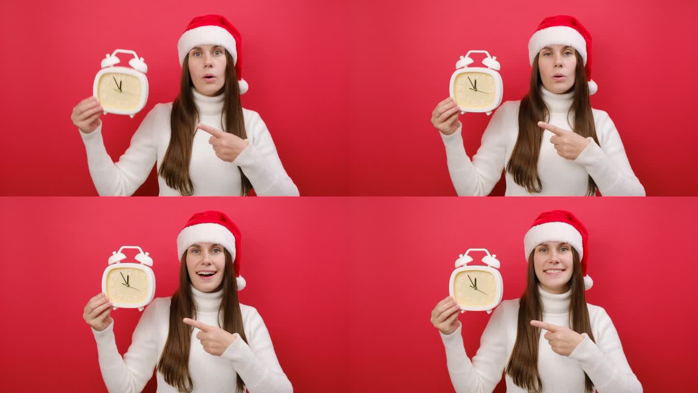 快乐的年轻女子穿着暖和的毛衣，戴着圣诞老人帽，用食指指着5点到12点的时钟，在淡红色的工作室背景下独