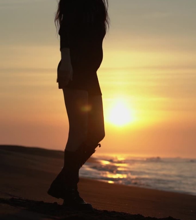 苗条的女人在日出时在沙滩上散步和转身。女士穿着夏日短裙和长靴