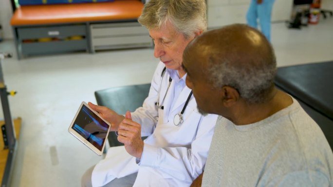 白人医生在医院一边看着黑人老人的x光片，一边向他解释受伤的原因