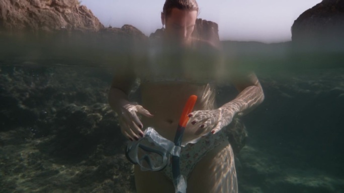 一个年轻女子站在清澈的海水中的岩石上，手里拿着一个带呼吸管的潜水面罩。