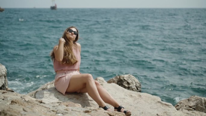 一个穿着粉色短裤的年轻女子坐在岩石上，背景是海浪和水花。她整理了一下头发