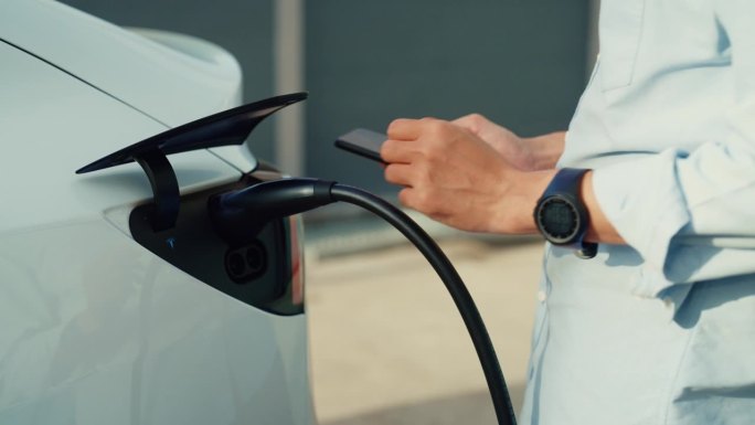 一名亚洲男子司机将电动汽车连接到电力系统，为汽车电池充电，并通过智能手机控制充电过程